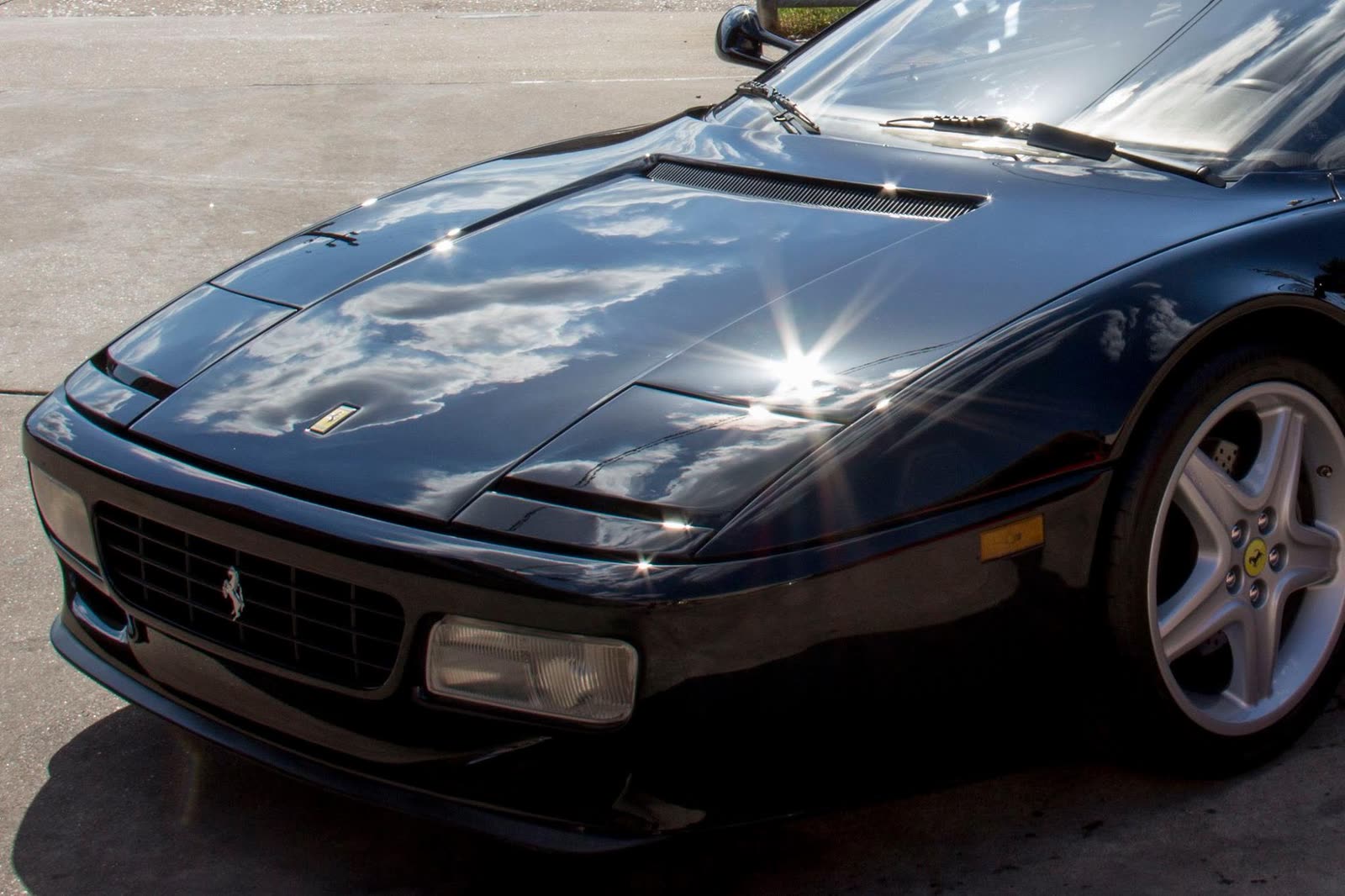 Clouds and the sun reflected in the black hood of a Ferrari 512 TR