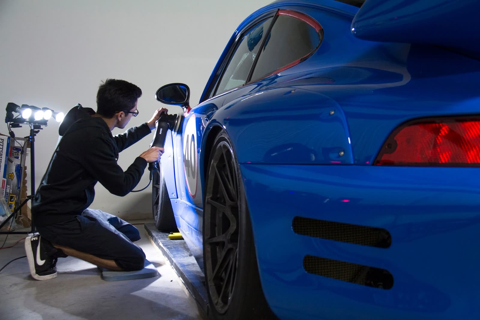 Kneeling to polish the door of a blue Porsche 911 beside a bank of work lights